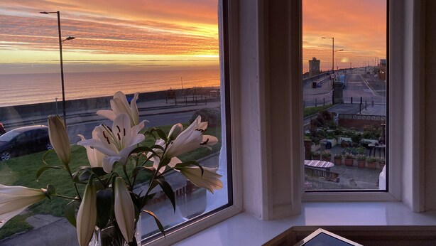 Une vue sur l’océan au coucher du soleil, prise à travers une fenêtre, avec un arrangement floral sur le rebord de la fenêtre.