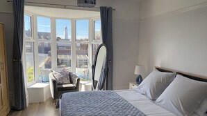 Une chambre avec un grand lit, une chaise, une table de chevet, un miroir et une vue sur les bâtiments à travers une grande baie vitrée.