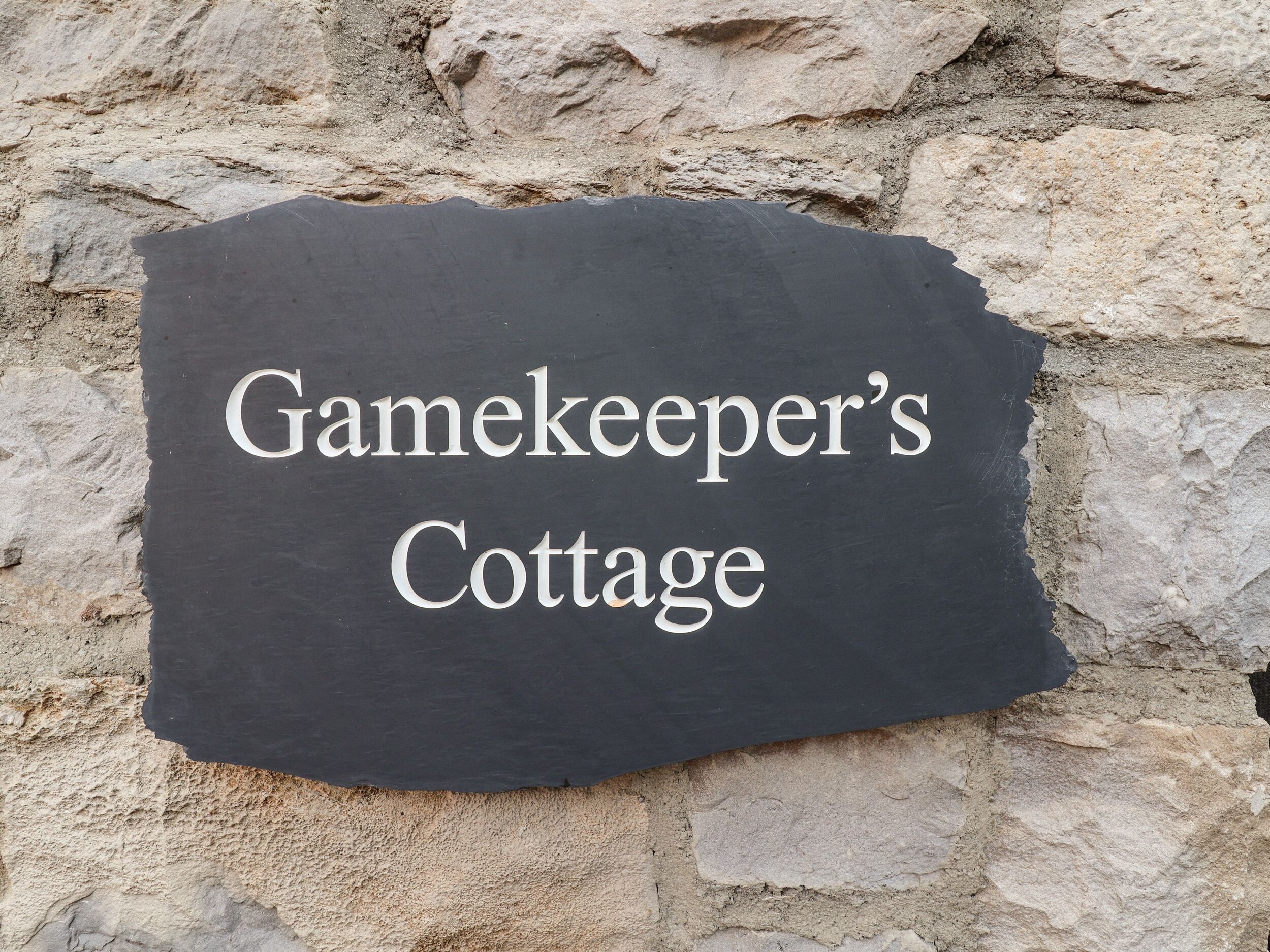 A slate sign with the text "Gamekeeper's Cottage" against a stone wall background.