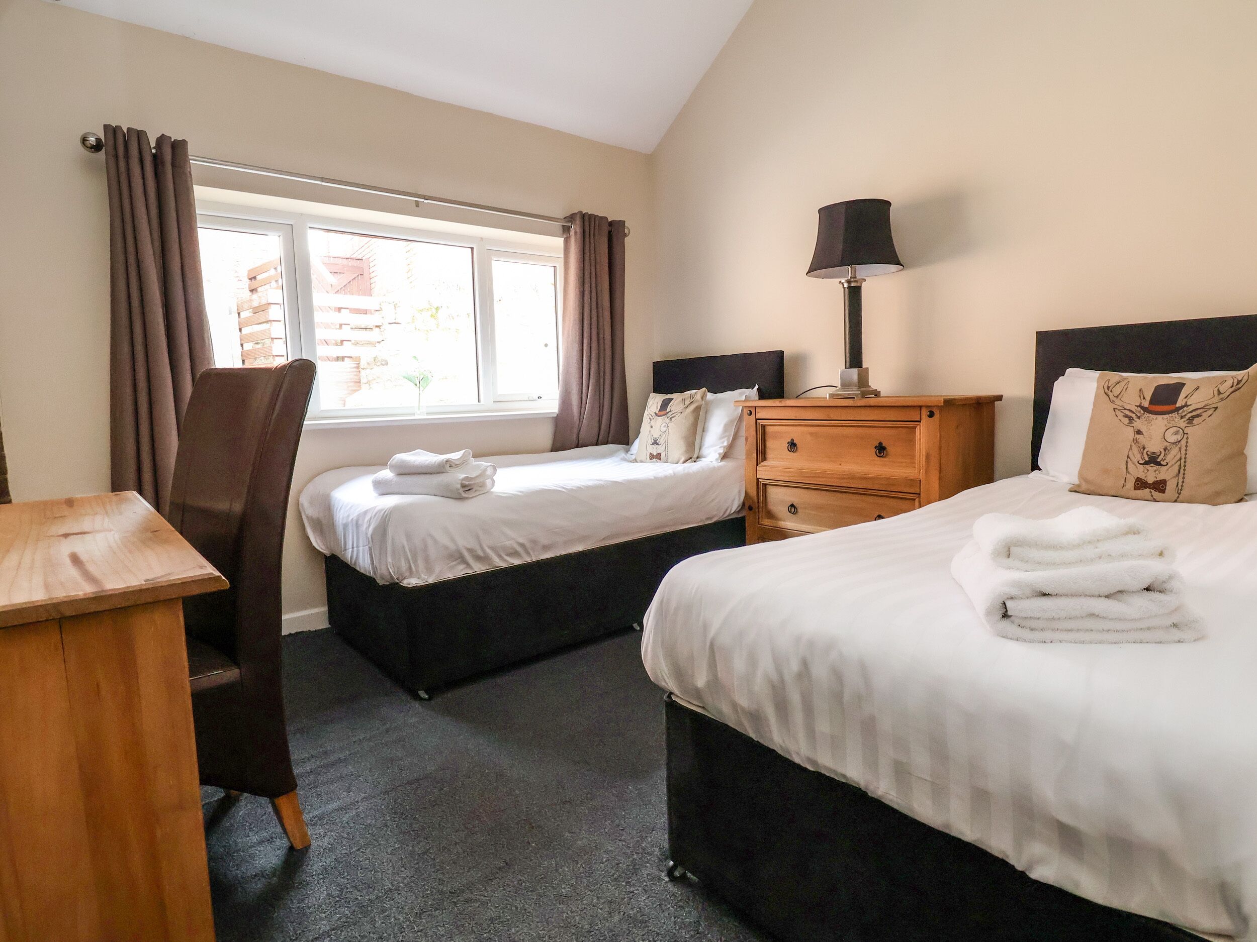 A hotel room with two beds, a wooden desk, a chair, a nightstand, and a window with curtains.
