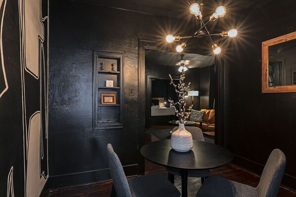 Una habitación oscura con una mesa redonda, sillas, un florero con ramas y una lámpara fijada a la pared.