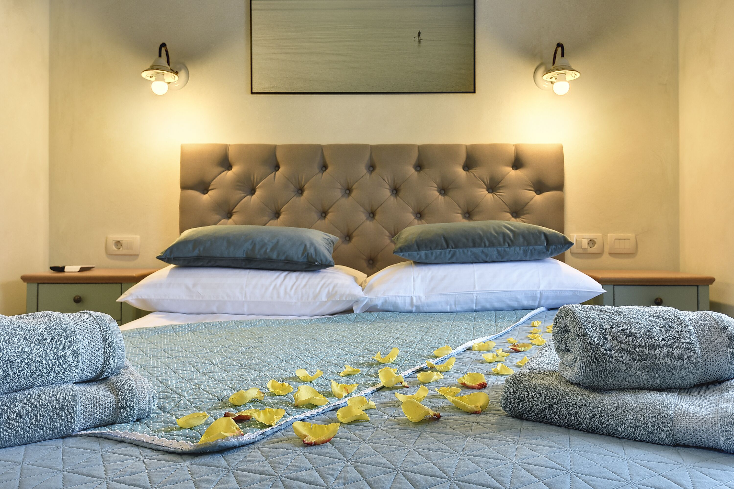 A bedroom with a tufted headboard, two bedside tables, a bed with a quilted blue bedspread, and two rolled towels.