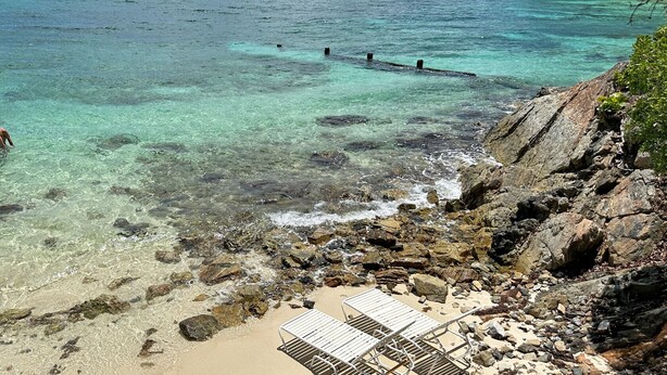 A rocky beach with clear water, a person in the water, and a metal bench.