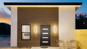 A modern building with a dark door, two illuminated wall sconces, and three wicker chairs outside.