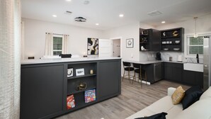 A modern kitchen with dark cabinetry, a white countertop, and a built-in island with shelves. There is a dining area with a white table and chairs, and a living room with a white sofa and a black throw pillow.