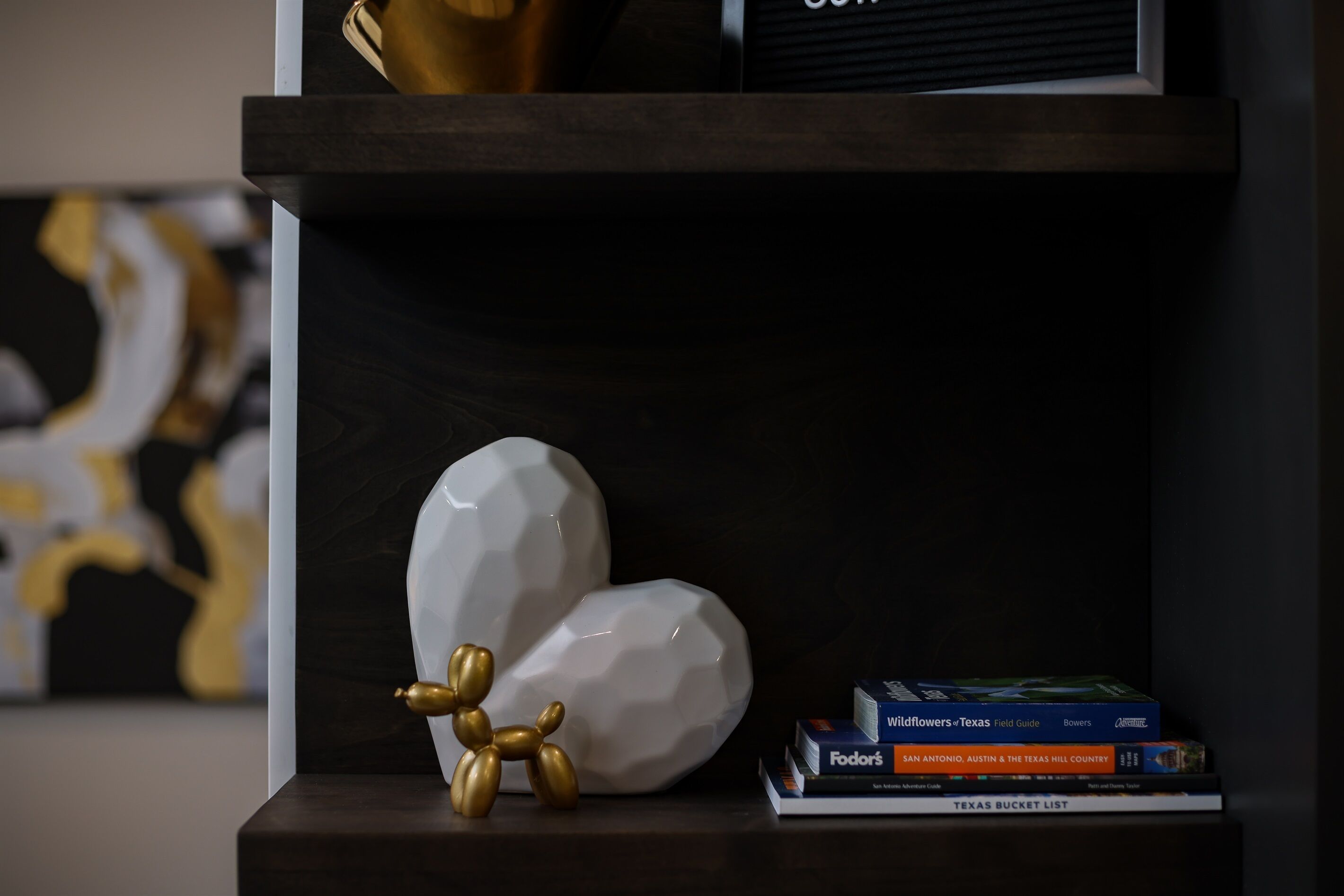 A shelf with a white geometric sculpture, a gold dog sculpture, and a stack of books including a Texas field guide.