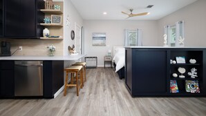 A modern kitchen with a dishwasher, wooden stools, a small dining table, and a built-in cabinet with decorative items. - Serene Hill Country Casita ~ Scenic View ~ Patio (Spring Branch)