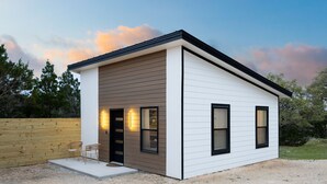 A modern single-story house with a flat roof, a front porch with two chairs, and a wooden fence in the background. - Serene Hill Country Casita ~ Scenic View ~ Patio (Spring Branch)