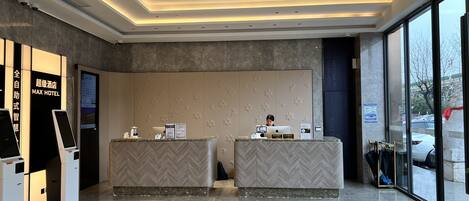 A modern hotel reception area with a digital kiosk, a reception desk, and a large window offering a view of the parking lot.