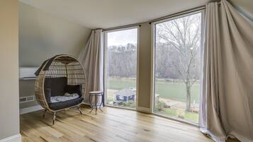 Une pièce dotée d'une grande fenêtre, d'un fauteuil en osier, d'une petite table et offrant une vue sur un lac et une maison.