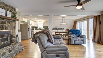 A modern living room with a stone fireplace, a ceiling fan, and a view into the kitchen.