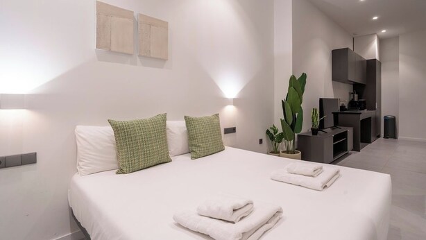 Ein modernes Hotelzimmer mit einem aufgeräumten Bett, einer kleinen Pflanze und einer Küchenzeile im Hintergrund.