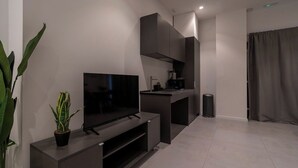 Ein modernes Wohnzimmer mit Flachbildfernseher auf einer dunklen Konsole, einer kleinen Küchenzeile mit Kaffeemaschine und einem Blumentopf.