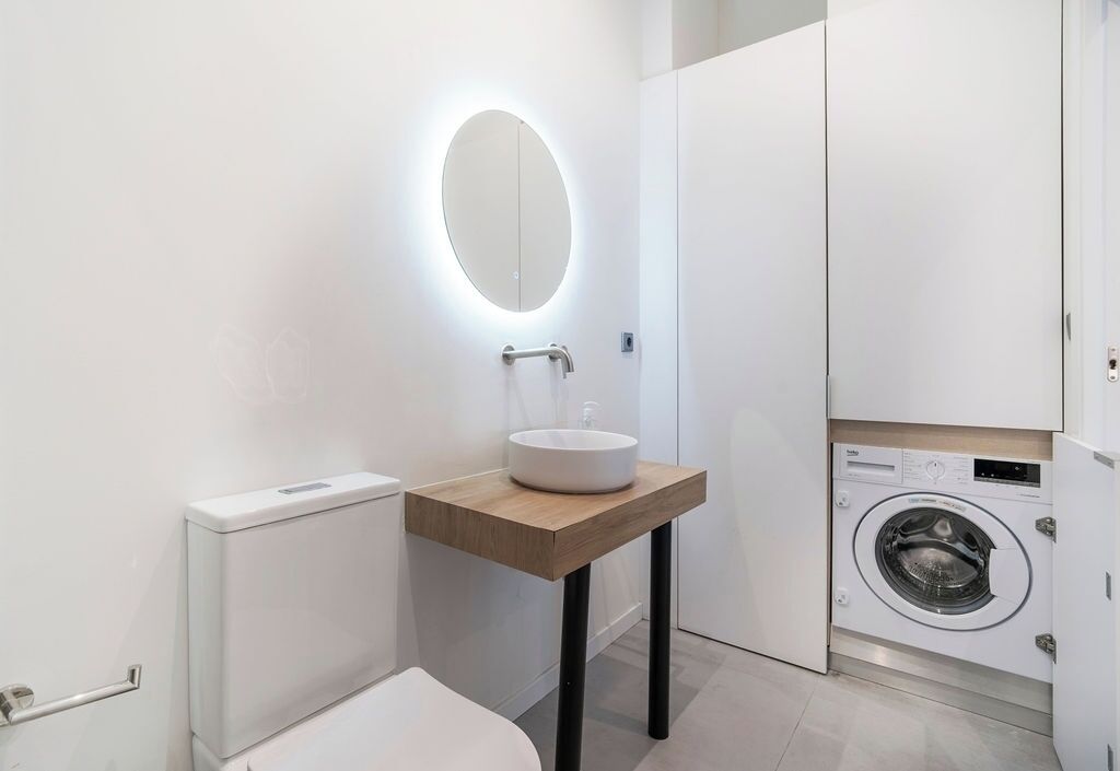 Ein modernes Badezimmer mit rundem Spiegel, Waschtisch aus Holz, Toilette und Waschmaschine.