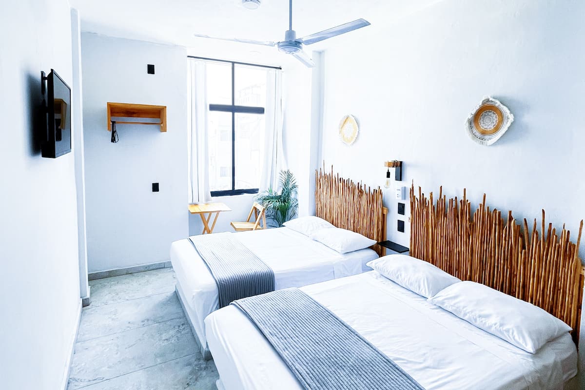 A hotel room with two beds, a ceiling fan, a small table, a chair, and a window with curtains.