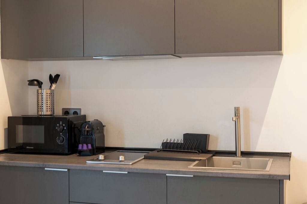 Une cuisine moderne avec des meubles gris, un four à micro-ondes encastré, une machine à café et un évier en acier inoxydable.