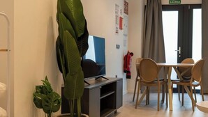 Een woonkamer met een televisie, een eettafel met stoelen, een kamerplant en een brandblusser.