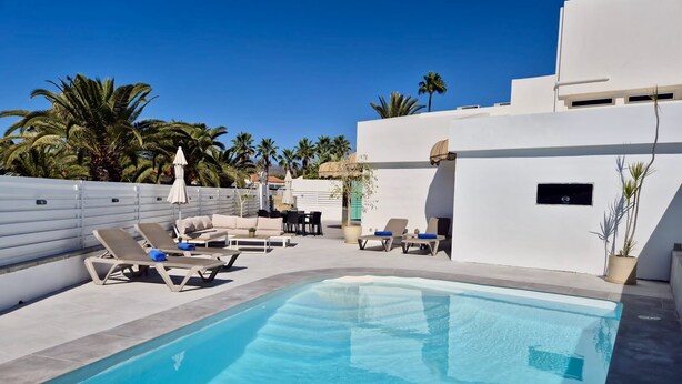 Un espace piscine extérieur moderne, doté de chaises longues et de parasols, entouré de palmiers et bordé d’un bâtiment blanc.