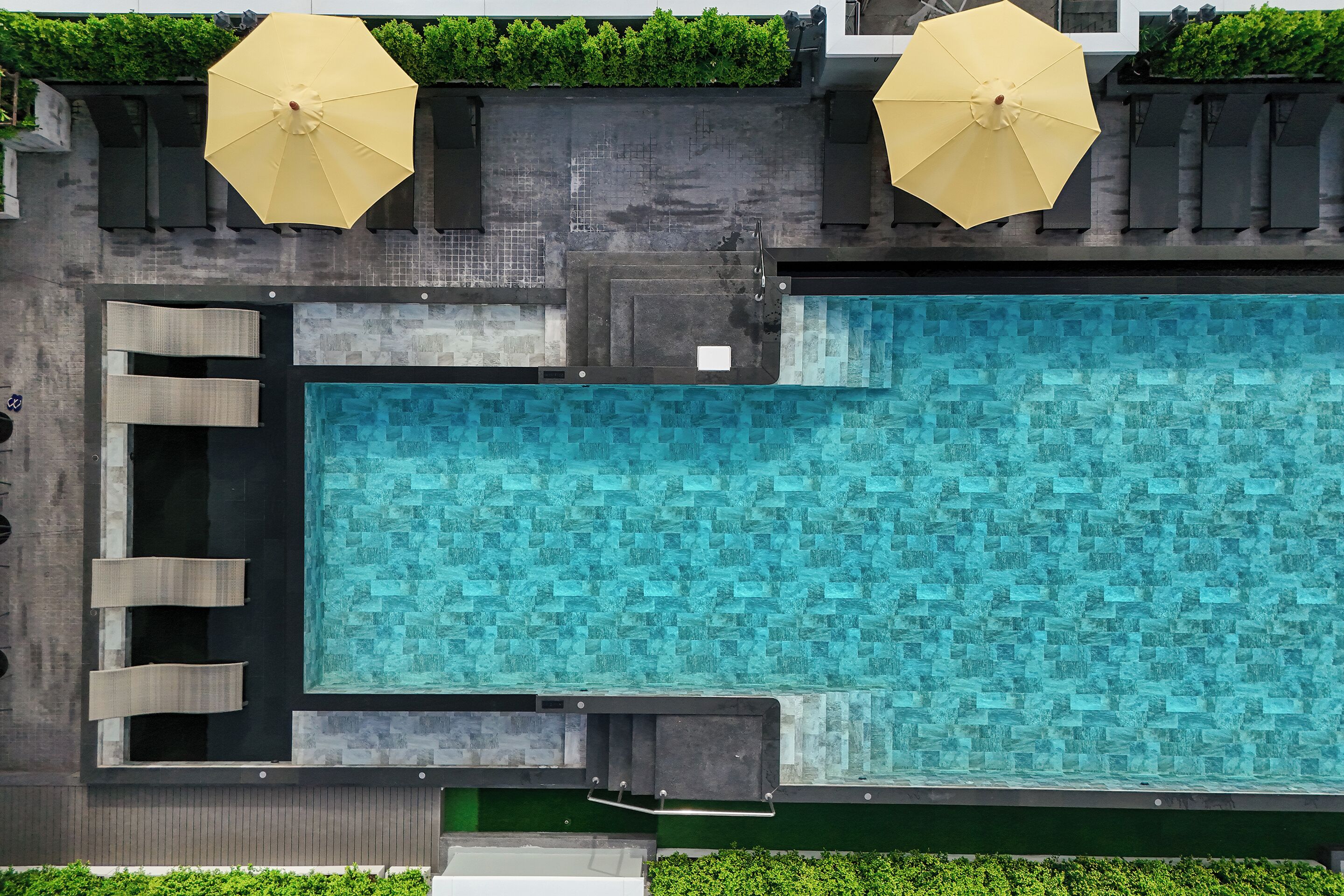 An aerial view of a rectangular swimming pool with a tiled deck, surrounded by greenery and two large umbrellas.