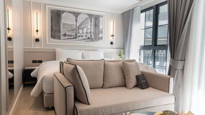A modern hotel room with a sofa bed, a dining table with breakfast, a framed picture on the wall, and a window with curtains. - Chateau Du Village Patong (Patong)