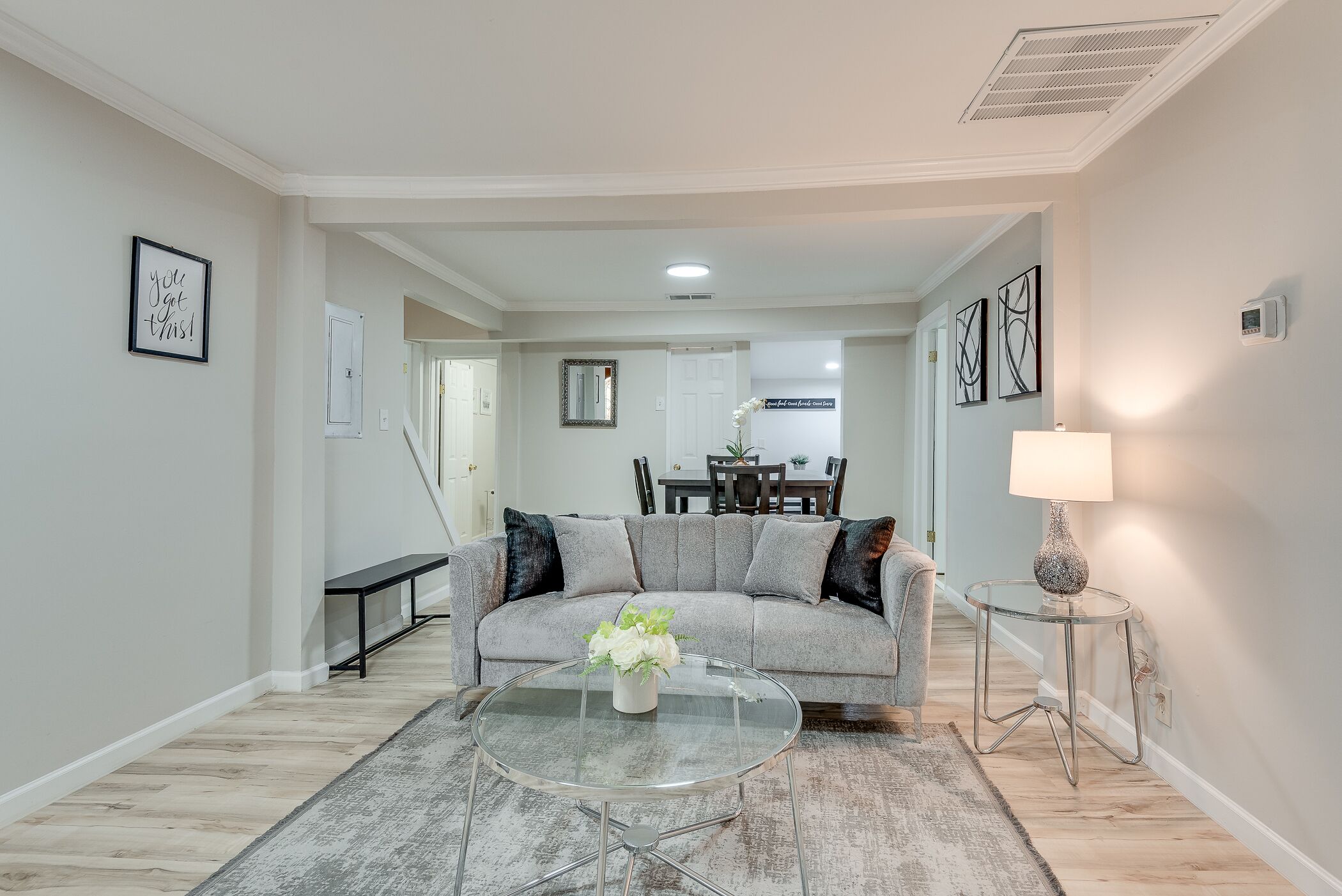 A living room with a grey sofa, a glass coffee table with a white vase, a dining area with a table and chairs, and a lamp on a side table.