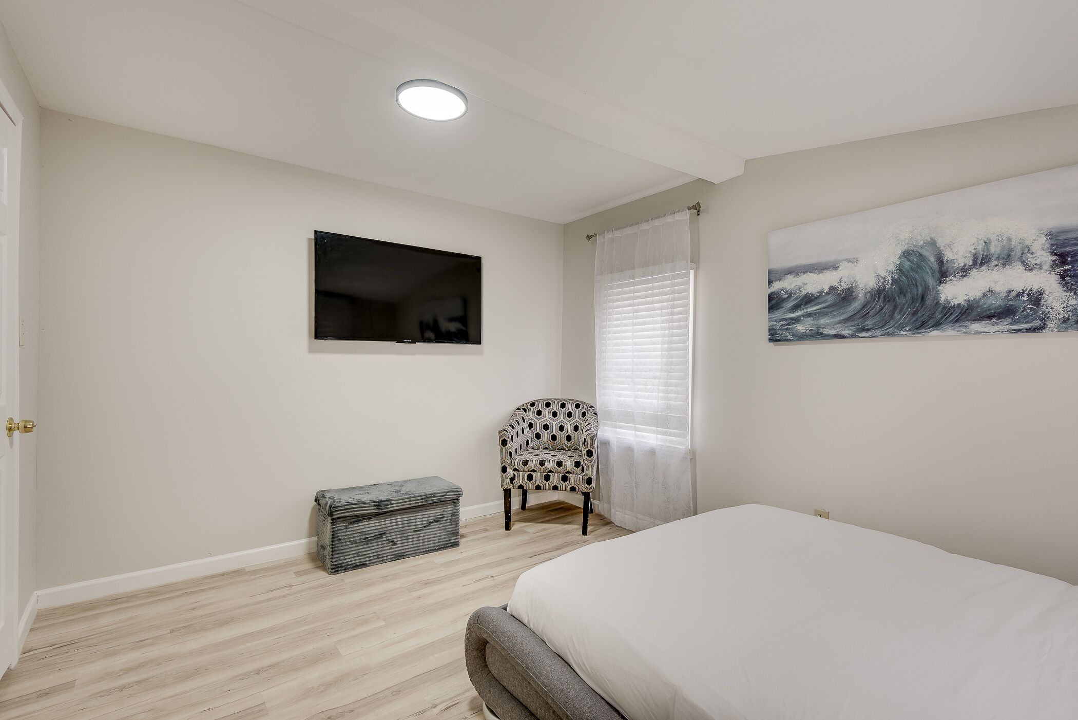 A bedroom with a bed, a chair, a small bench, a painting, and a ceiling light.