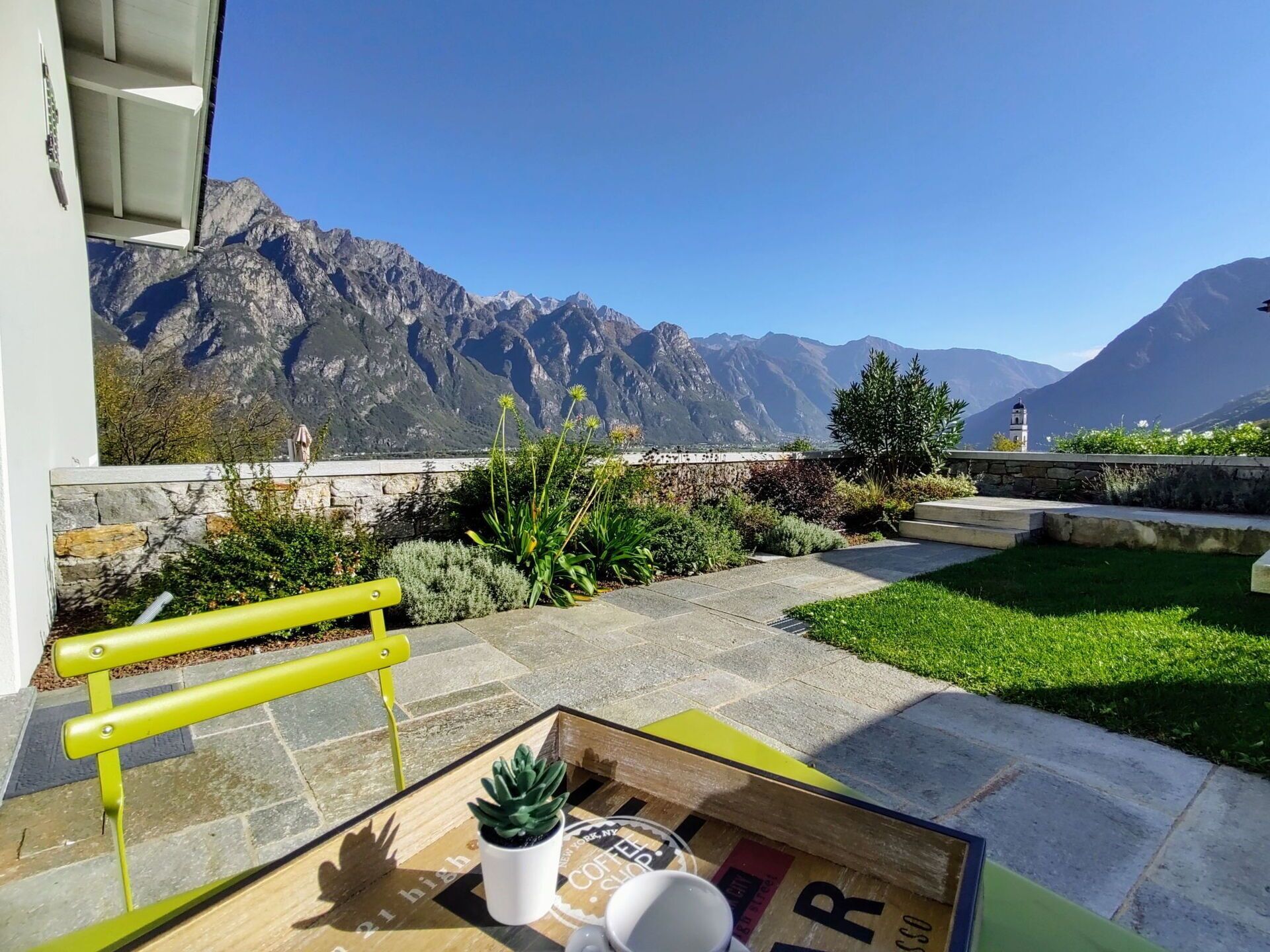 A terrace with a view of mountains, a yellow bench, a table with a potted plant, and a coffee cup.