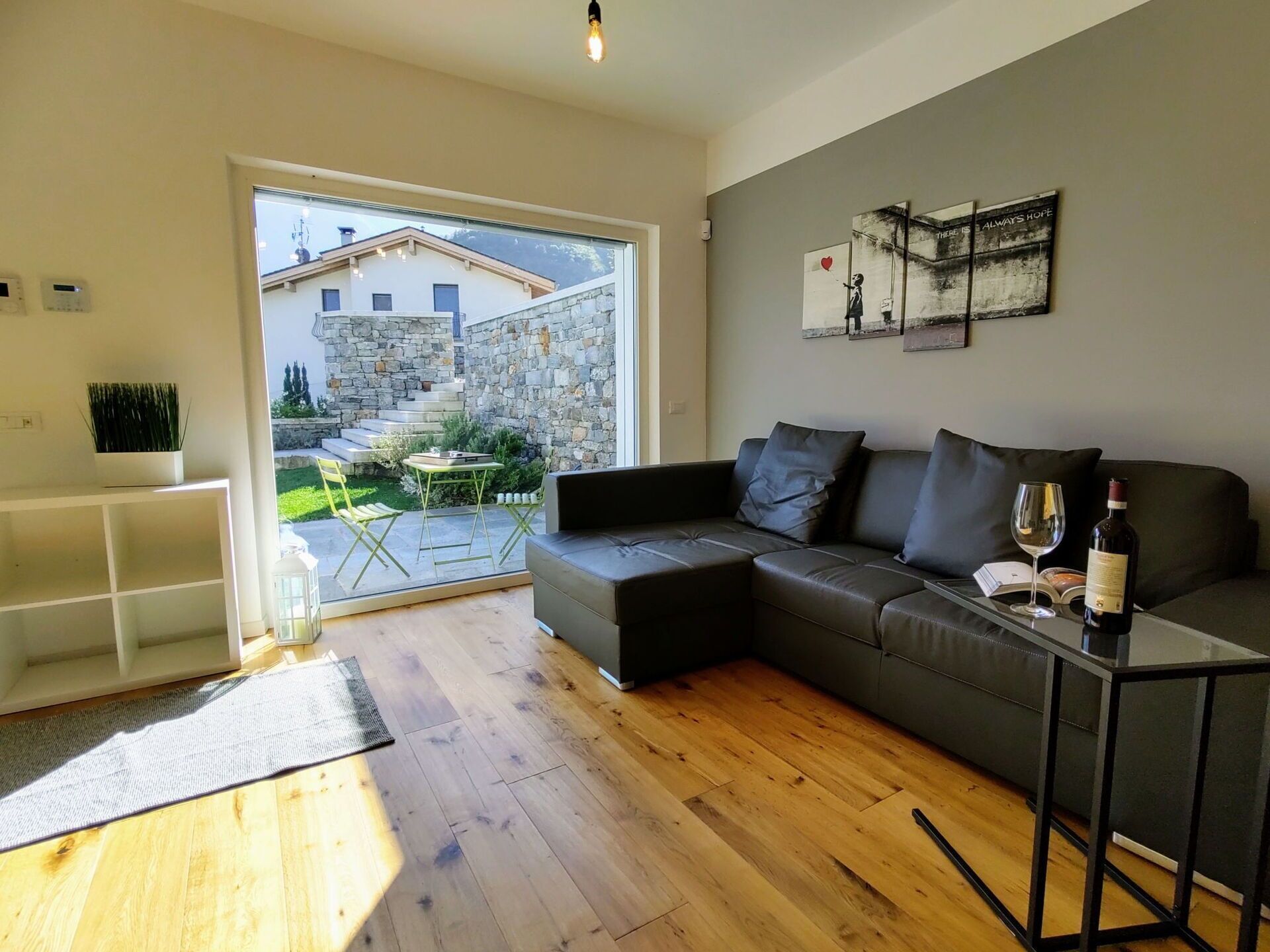Un salon moderne avec un canapé gris, une table basse et une bouteille de vin. On aperçoit, à travers une porte vitrée, un bâtiment en pierre et un jardin.