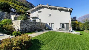 Une maison moderne à deux étages, dotée d’un mur en pierre, d’une pelouse bien entretenue et offrant une vue sur les montagnes.