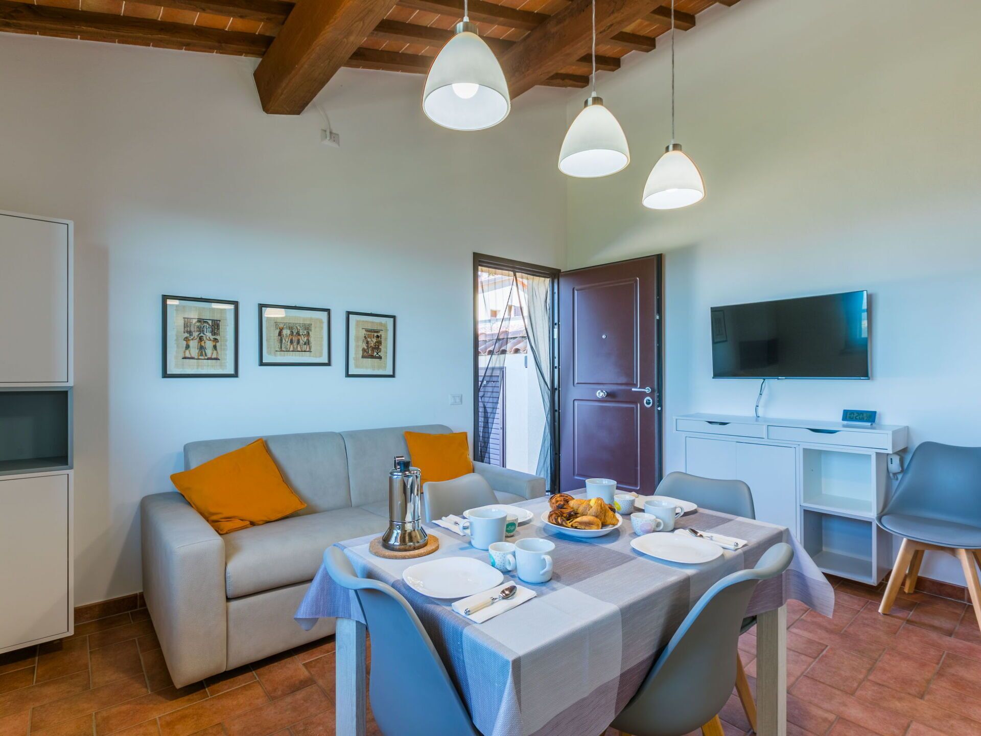 Un coin repas avec une table dressée pour deux personnes, un canapé avec des coussins orange et un téléviseur fixé au mur.