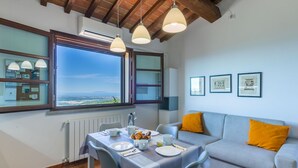 A dining area with a table set for two, a sofa with a yellow pillow, and a window with a view of the landscape.