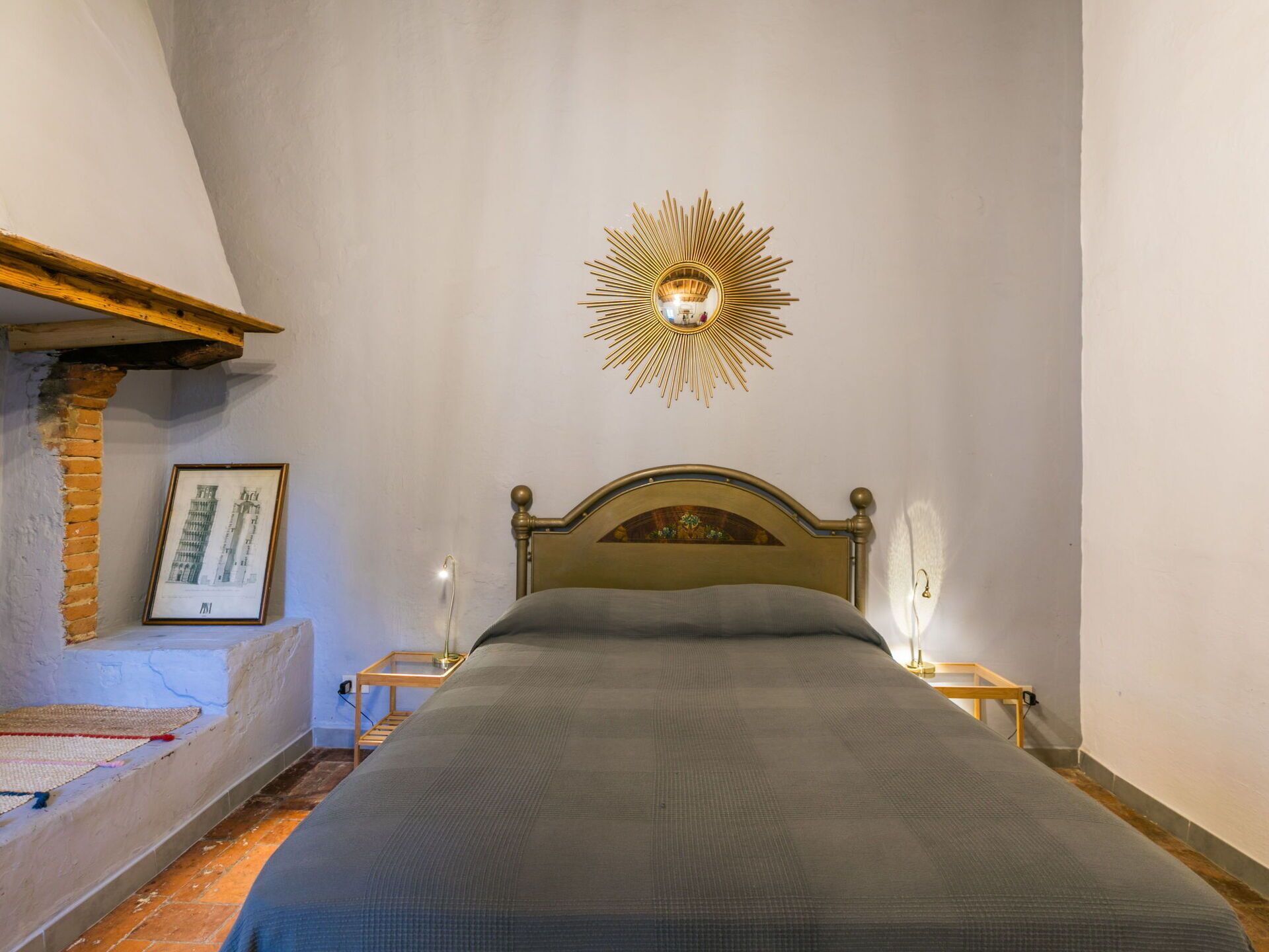 A bedroom with a bed, a wall-mounted sunburst mirror, a framed architectural drawing, and a wooden nightstand with a lamp.