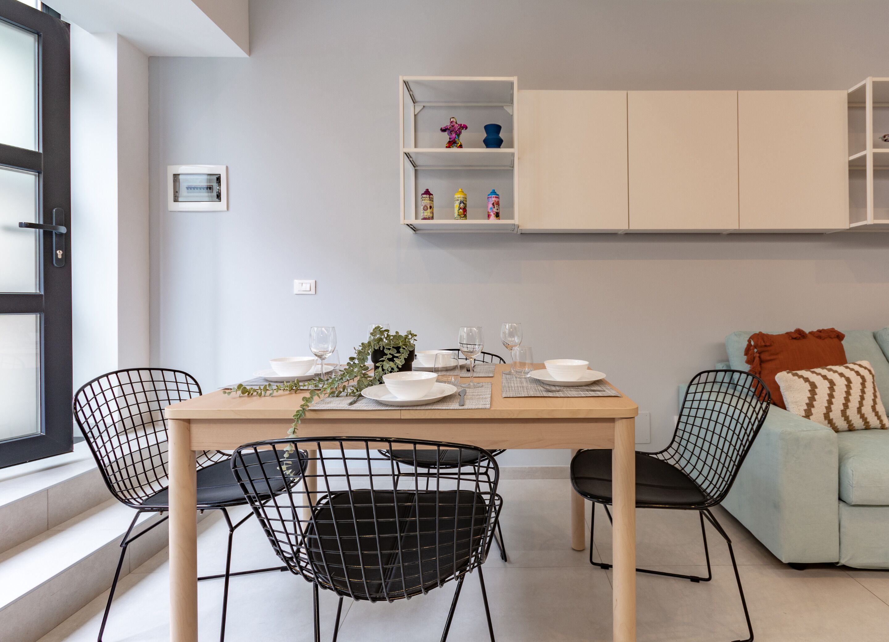 Un comedor moderno con una mesa dispuesta para cuatro personas, rodeada de sillas con estructura de alambre negro. Un sofá de color claro con cojines estampados está apoyado en la pared, y un armario blanco con estantes se encuentra sobre la mesa.