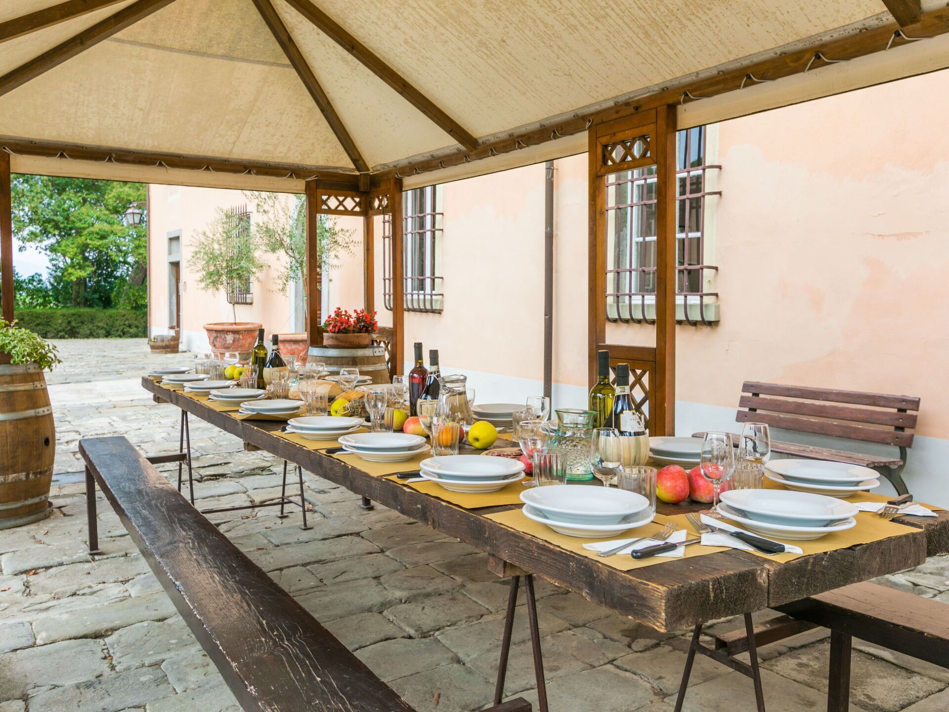 A long dining table set outdoors under a canopy, with plates, glasses, and bottles of wine.