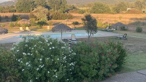 A pool area with lounge chairs and thatched umbrellas, surrounded by greenery and hills in the background.