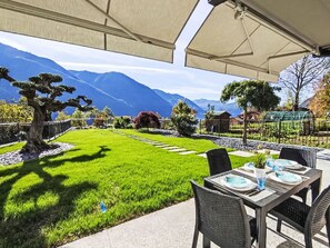 An outdoor patio with a dining table set for two, a large tree, and a mountain view.