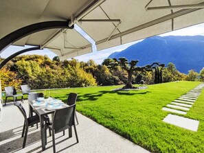 Ein überdachter Außenbereich mit Tisch und Stühlen, einem gepflegten Rasen und Blick auf ein Gebirge.