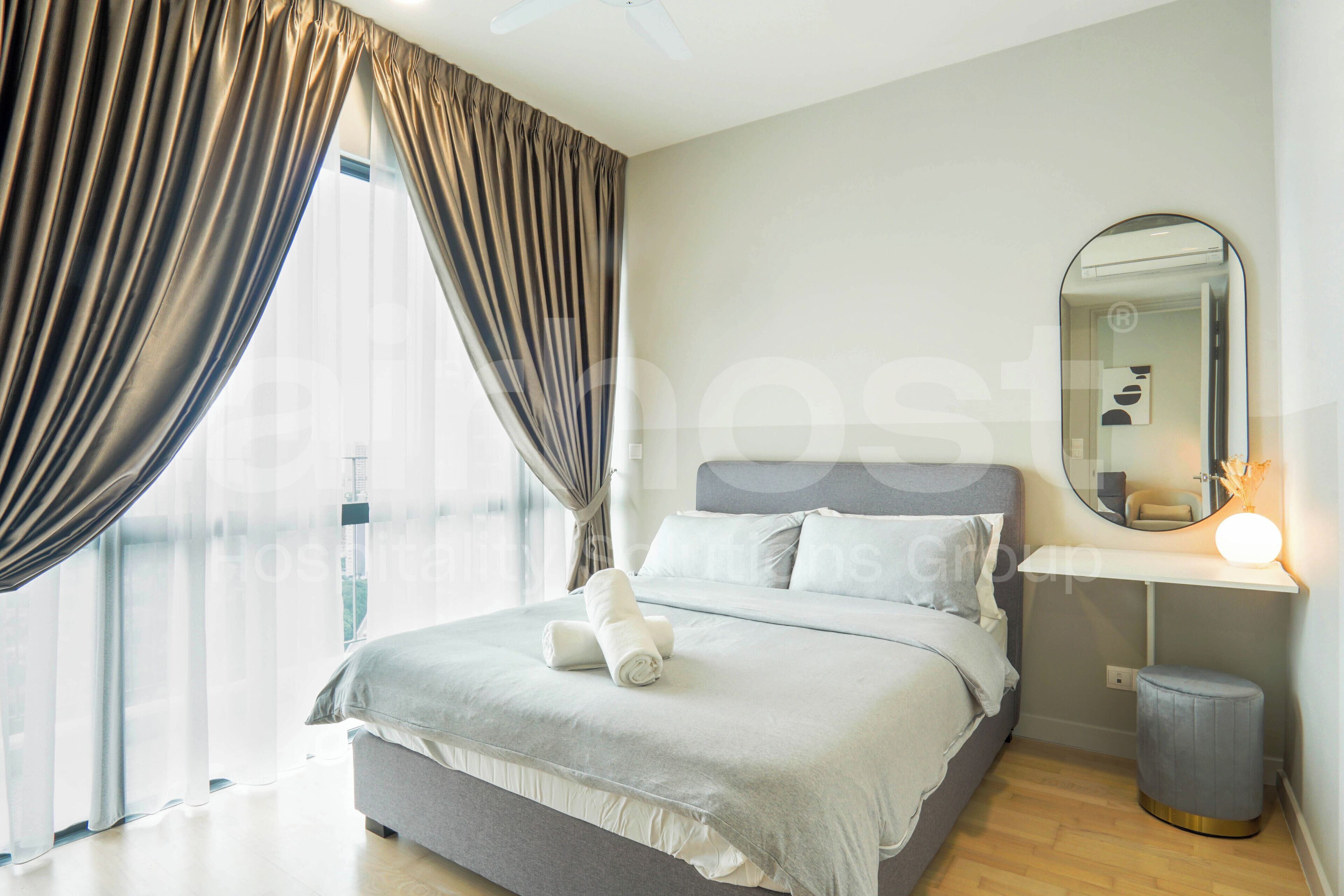 Habitación de hotel moderna con una cama grande, una mesita de noche con lámpara, un espejo y vistas a la ciudad a través de cortinas translúcidas.