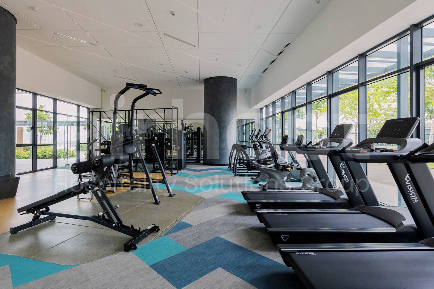 A modern gym with cardio machines, weight training equipment, and large windows offering views of the outdoors.