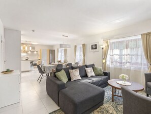 Uma sala de estar moderna com um sofá seccional cinza, uma área de jantar com mesa e cadeiras, e uma cozinha ao fundo.