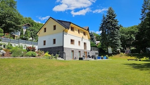 Une maison à deux étages, avec une base en pierre et un étage supérieur jaune, entourée d’une pelouse bien entretenue et d’arbres.