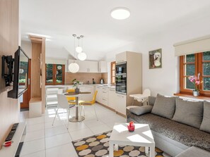 Una cuina moderna amb armaris blancs, una zona de menjador amb cadires grogues, un sofà blanc i un televisor fixat a la paret.