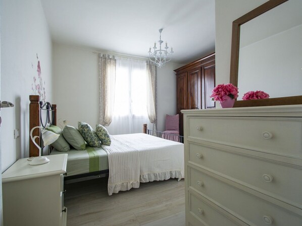 A bedroom with a bed, a dresser, a chair, a window with curtains, and a chandelier.