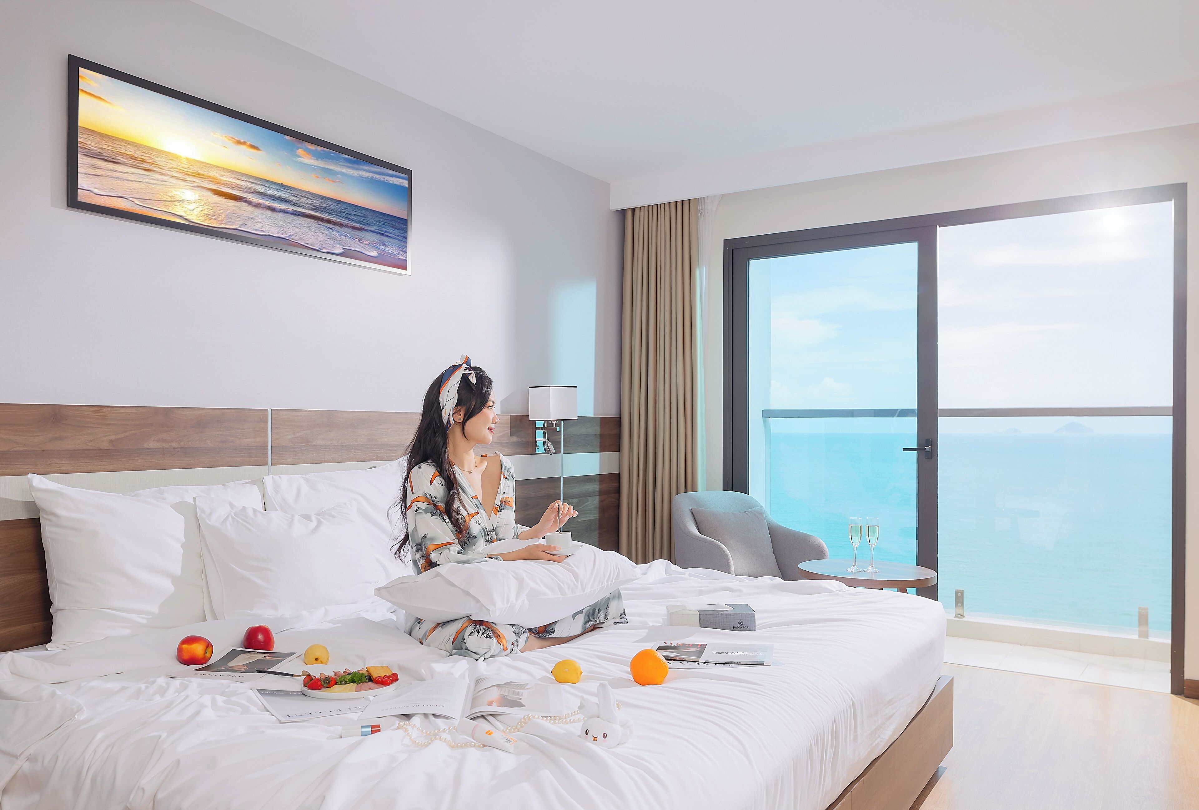 A hotel room with a large bed, a view of the sea, a painting on the wall, and a table with two glasses.