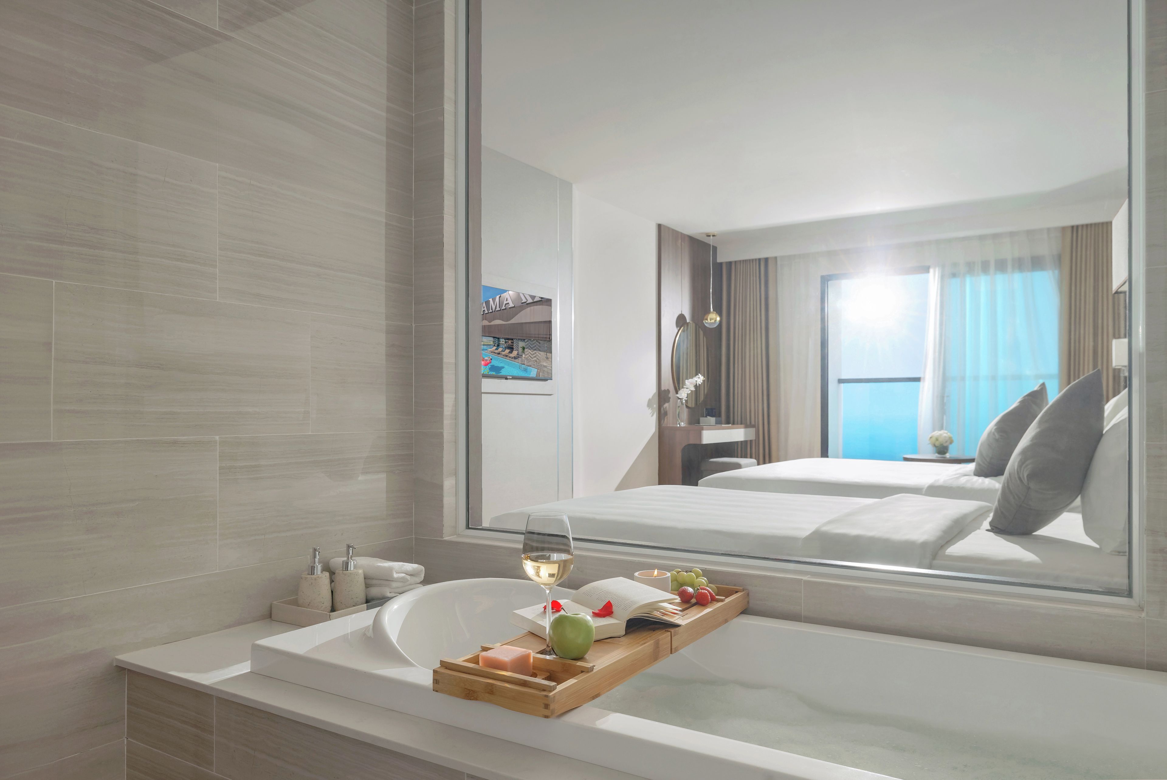 Habitación de hotel moderna con un espejo grande, una bañera con una bandeja de frutas y un libro, y vistas al área de la piscina.