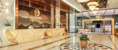 A modern hotel lobby with a reception desk, a large chandelier, and a decorative floor pattern.