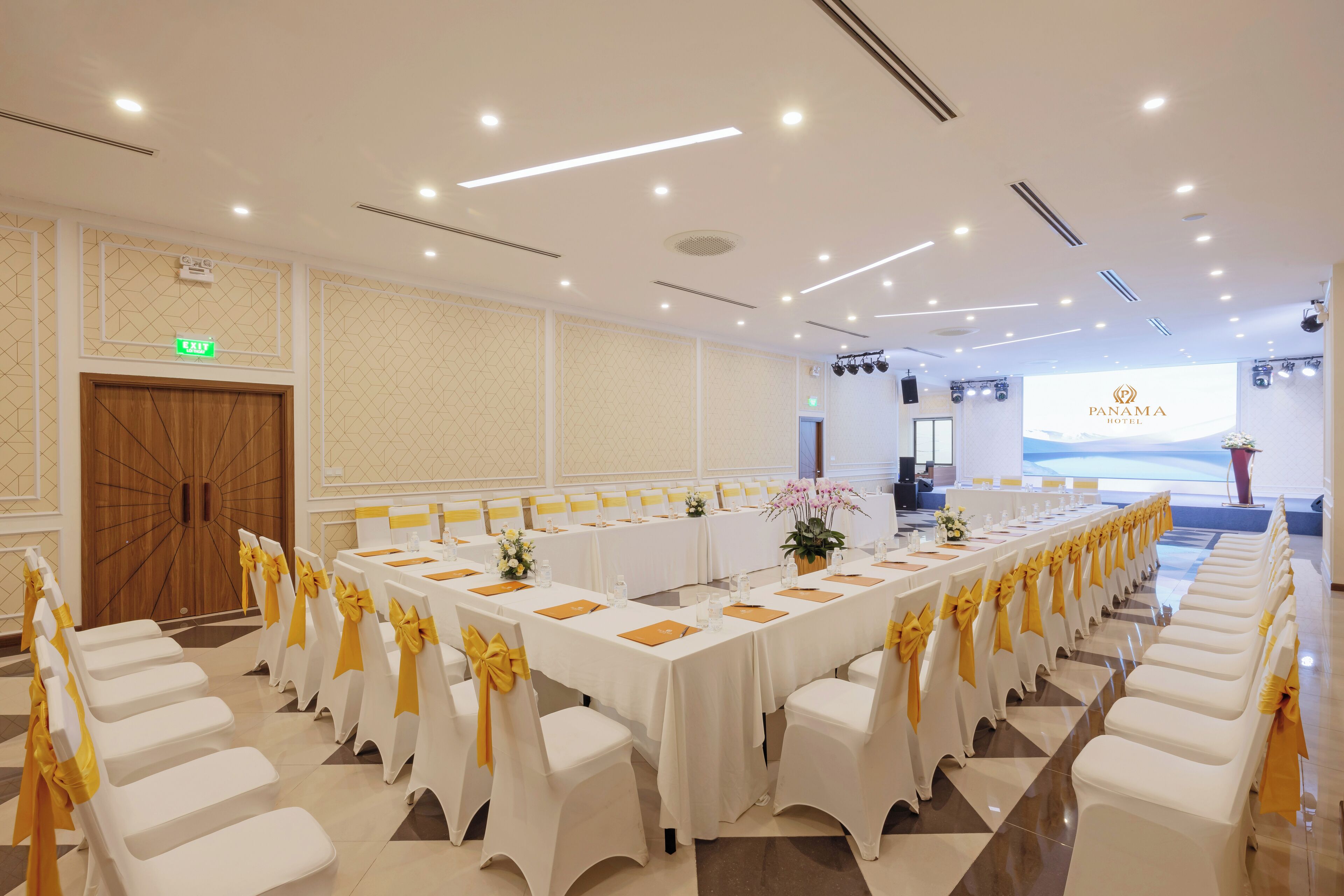 A conference room with white tables and chairs, yellow sashes, and a large screen displaying the Panama Hotel logo.
