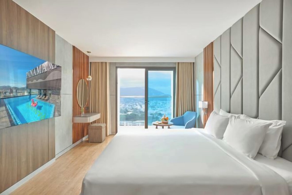 A hotel room with a large bed, a TV mounted on the wall, a view of the city, and a balcony.