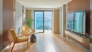 Een moderne hotelkamer met een grote televisie, een zithoek met een tafel en stoelen, en een balkon met uitzicht op de stad.
