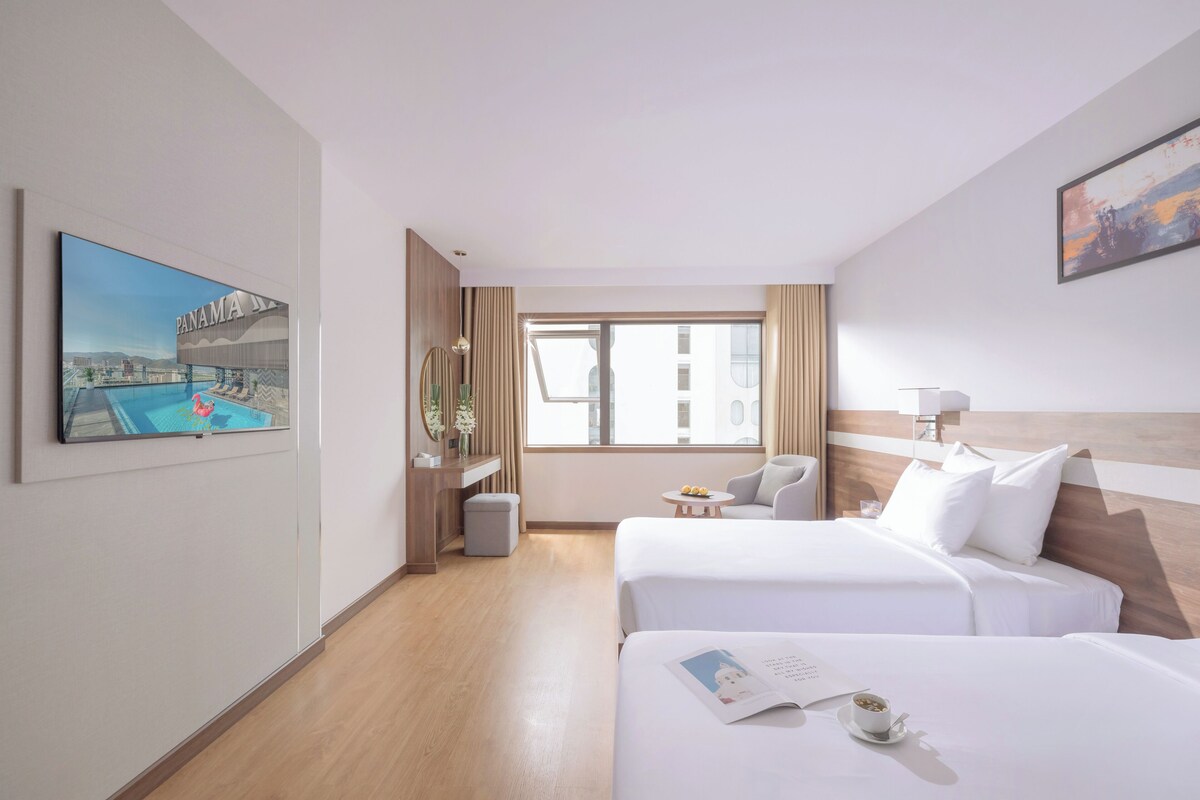 Une chambre d’hôtel avec deux lits, une télévision, une fenêtre donnant sur un beau paysage et une petite table avec une lampe.